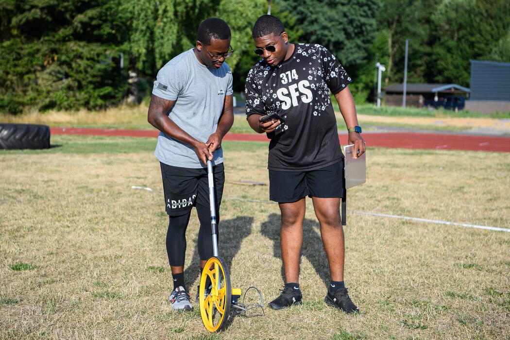 Two men look at a phone while one holds a measuring wheel on the ground.