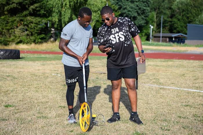Two men look at a phone while one holds a measuring wheel on the ground.