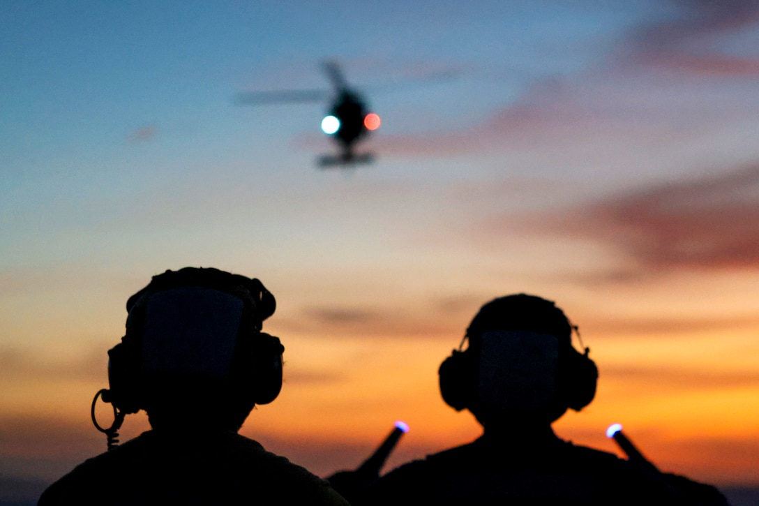 Two sailors, one holding a light device in each hand, watch as a helicopter descends, with an orange and blue sky in the background.
