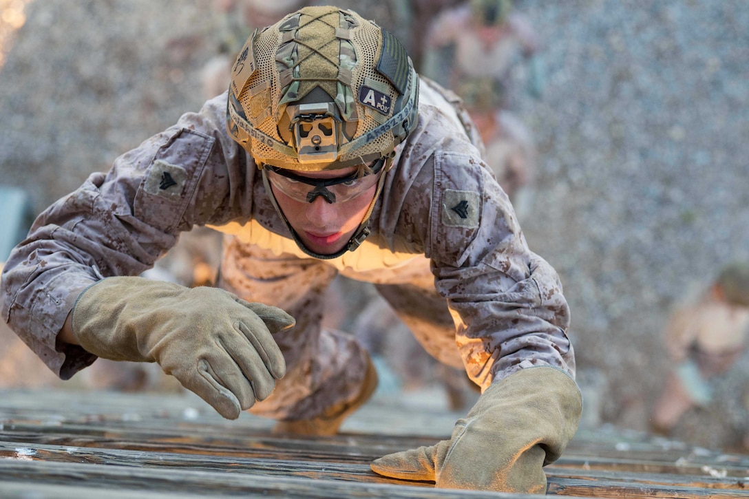 A Marine wearing heavy gloves and a helmet looks up and climbs a wooden tower as others stand below.