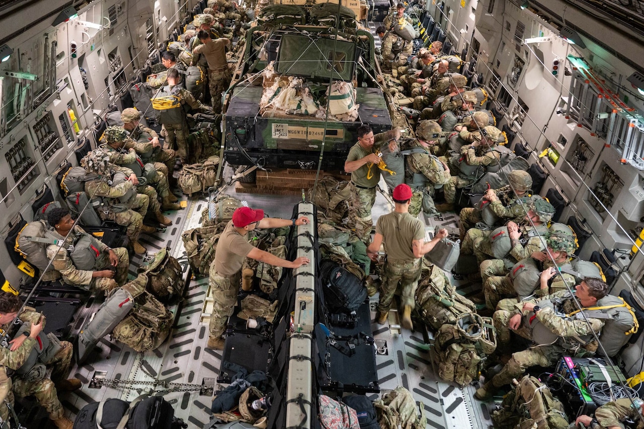 Dozens of troops in camouflage uniforms wearing parachutes sit inside an aircraft.