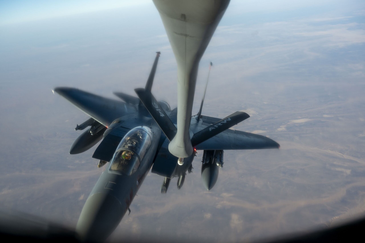 A nozzle extends down toward a jet flying high above a hazy terrain.