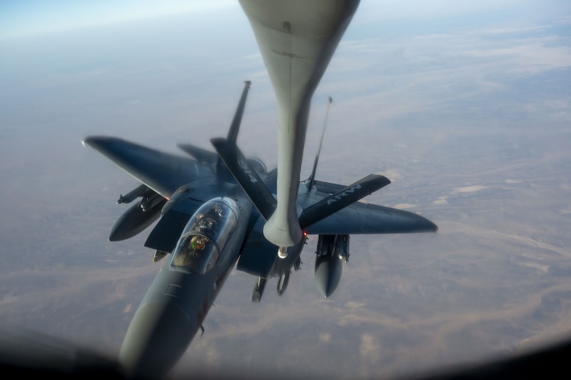 A nozzle extends down toward a jet flying high above a hazy terrain.
