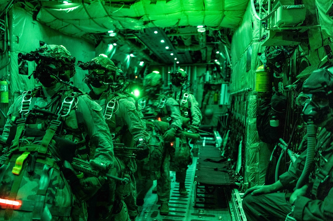 Marines in parachute gear stand in a line inside a green-lit aircraft with a person seated to the side.