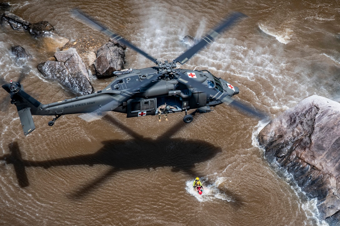 A soldier sits forward from the open door of a helicopter hovering above a body of water and holds a line attached to a rescue basket with two people in it in the water below.