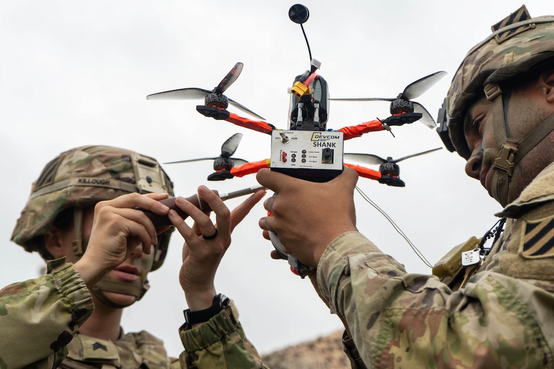 Two soldiers, one using a screwdriver, work on a small drone with propellers outdoors.