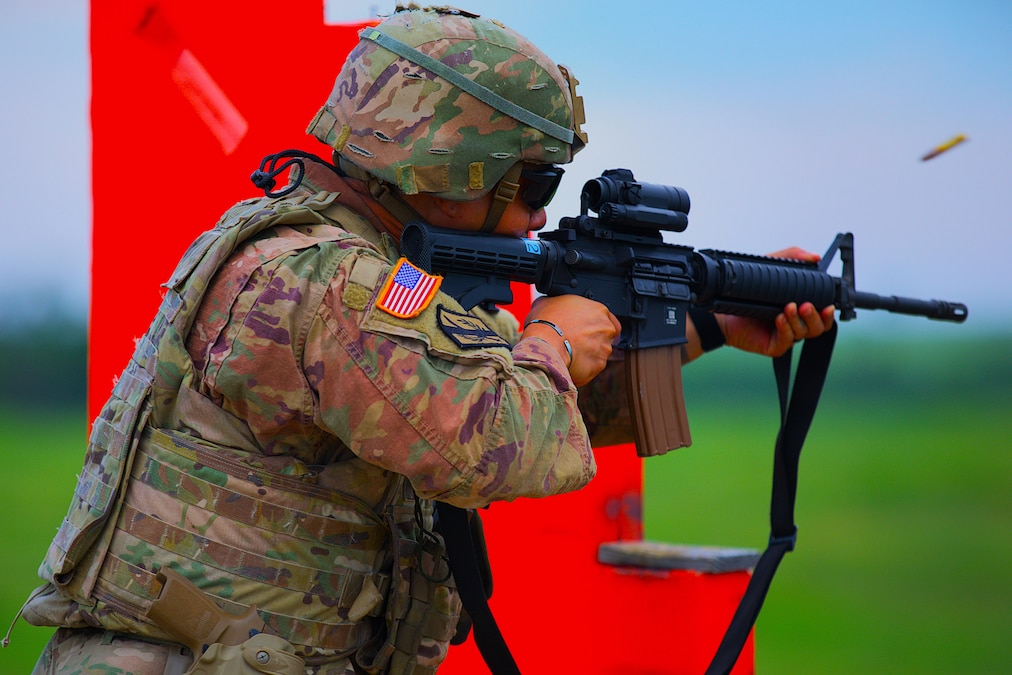 A soldier standing near a bright red obstacle fires a rifle at an outdoor range.
