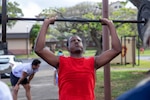 Service member wearing red tank top lifts weights outdoors.
