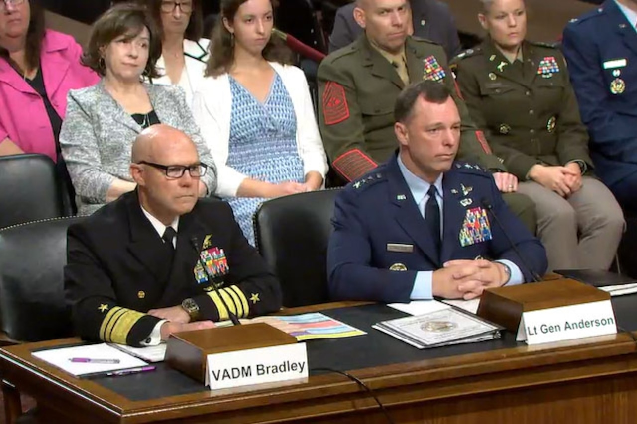 Two men in formal military uniforms sit at a long wooden desk while people sit behind them.