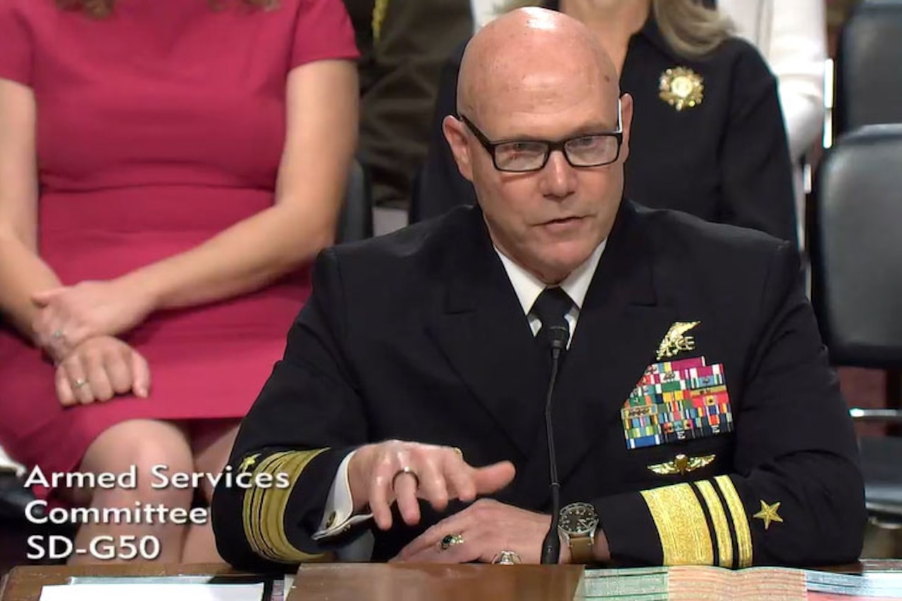 A man wearing a formal military uniform talks while seated behind a large wooden desk.
