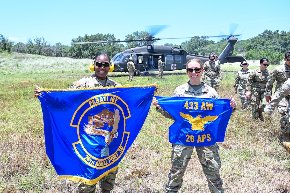 Master Sgt. Thea Lau, 26th Aerial Port Squadron Readiness and Resources flight chief, and Tech. Sgt. Tamekia Alston, 74th Aerial Port Squadron unit deployment manager holds up their respective unit flags while their fellow Airmen trained in a simulated Army medevac during OPERATION DUSTOFF VIGILANCE ’25 at Joint Base San Antonio-Camp Bullis, Texas, July 16, 2025.