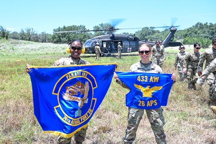 Master Sgt. Thea Lau, 26th Aerial Port Squadron Readiness and Resources flight chief, and Tech. Sgt. Tamekia Alston, 74th Aerial Port Squadron unit deployment manager holds up their respective unit flags while their fellow Airmen trained in a simulated Army medevac during OPERATION DUSTOFF VIGILANCE ’25 at Joint Base San Antonio-Camp Bullis, Texas, July 16, 2025.