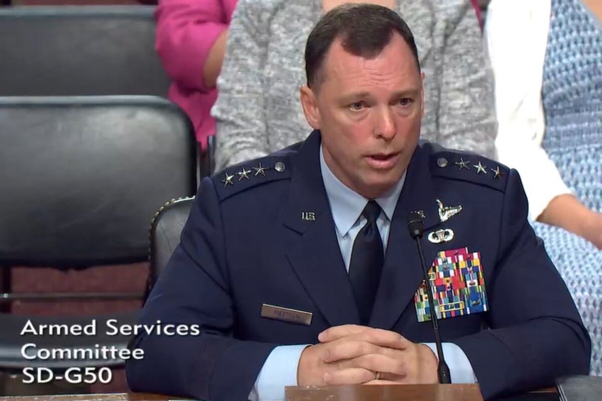A man wearing a formal military uniform talks while seated behind a large wooden desk.