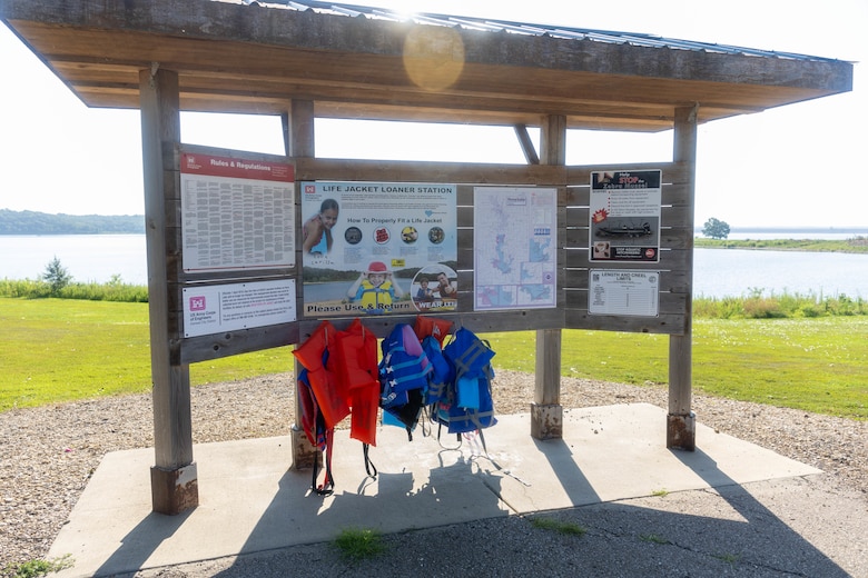 A wooden board with a wooden roof displays white posters and orange and blue lifejackets with grass and a lake in the background.