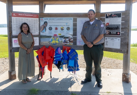 A woman in a green dress and a man in a grey shirt and green pants stand and smile in front of a wooden board with white posters and orange and blue lifejackets hanging in the background.