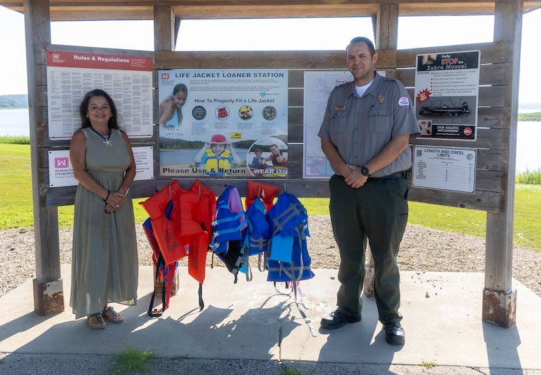 A woman in a green dress and a man in a grey shirt and green pants stand and smile in front of a wooden board with white posters and orange and blue lifejackets hanging in the background.