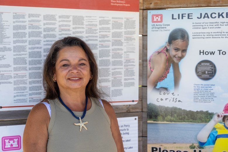 A woman in a green shirt stands and smiles in front of a wooden board with white posters hanging in the background.