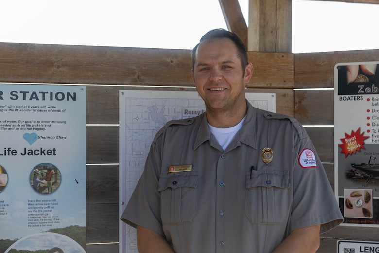 A man in a grey shirt stands and smiles in front of a wooden board with white posters hanging in the background.