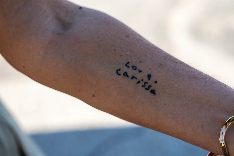 A woman's arm has a small black tattoo with the words "Love, Carissa".