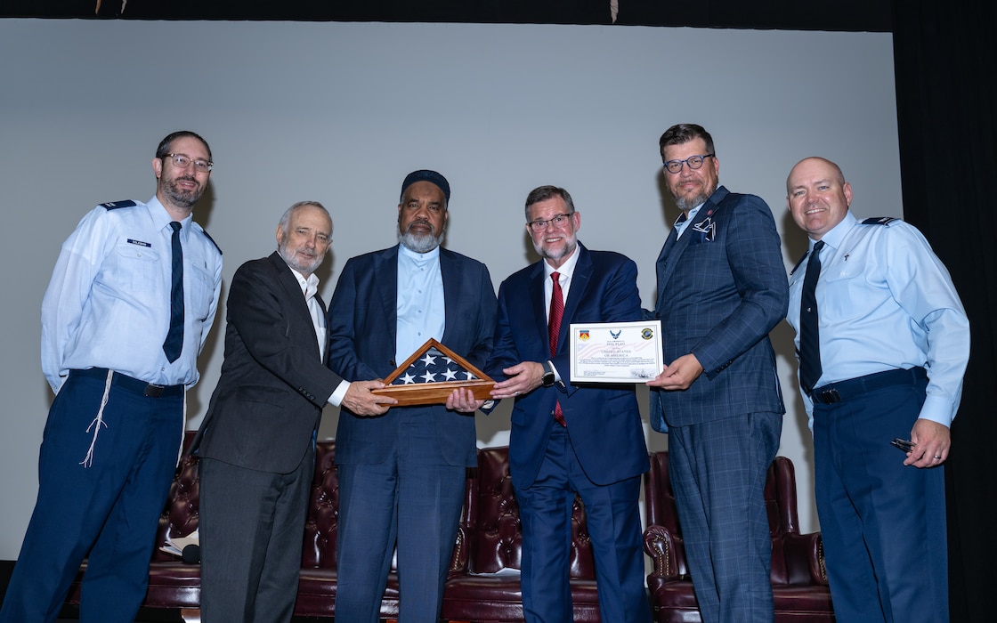 Two men in blues military uniforms stand on either side of a group of four men in suits holding a folded U.S. flag in a shadow box and a certificate