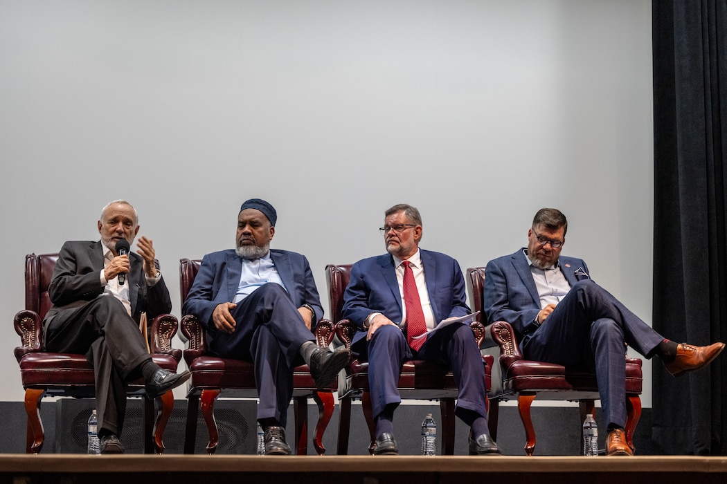 Four men in suits sit on stage, the man on the far left speaks into a microphone