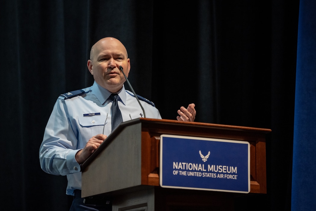A man in military blues uniform, stands behind a microphone and podium with a sign that reads national museum of the U.S. Air Force