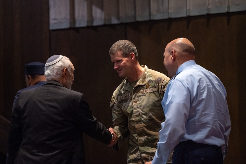A man in a camouflage military uniform shakes hands with a man in dark suit and yamaka as a man in a light blue shirt stands in front