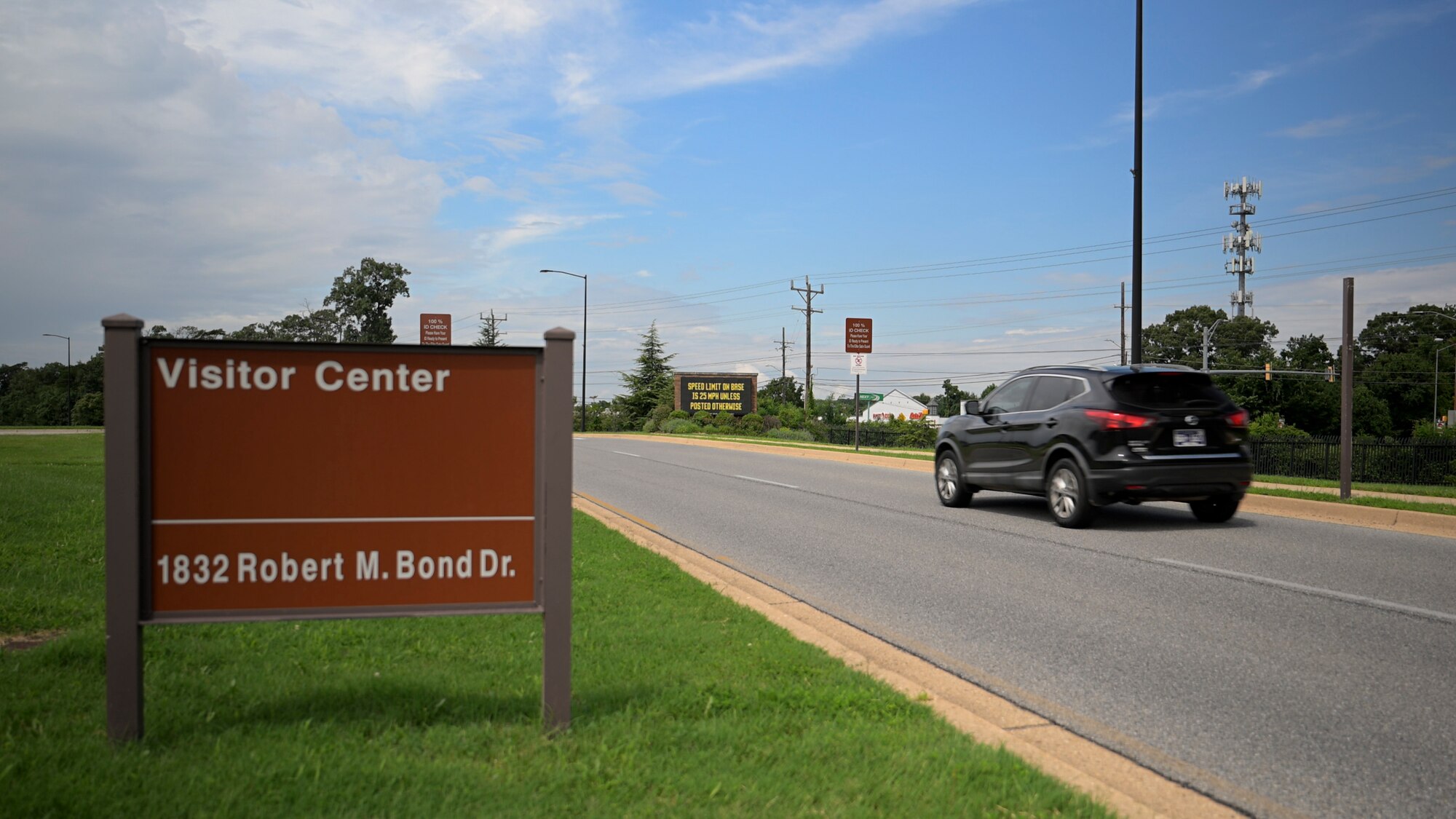 A black SUV drives on a road past a brown sign reading “Visitor Center, 1832 Robert M. Bond Dr.” near Joint Base Andrews. The area includes grassy shoulders, utility poles, and clear skies.