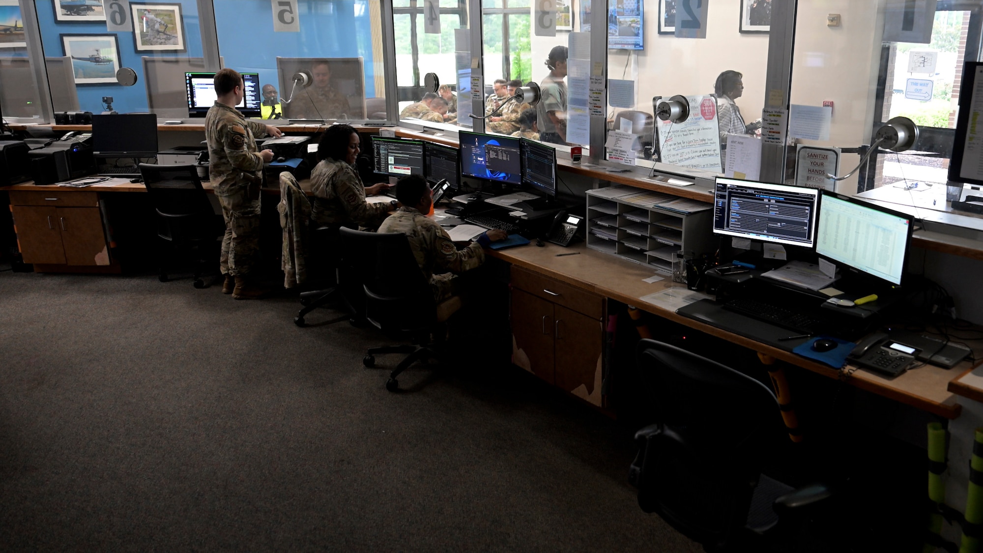 Several U.S. Air Force personnel in uniform work at computer stations inside a visitor control center. The room contains multiple monitors, desks, and windows looking into adjacent areas.