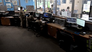 Several U.S. Air Force personnel in uniform work at computer stations inside a visitor control center. The room contains multiple monitors, desks, and windows looking into adjacent areas.