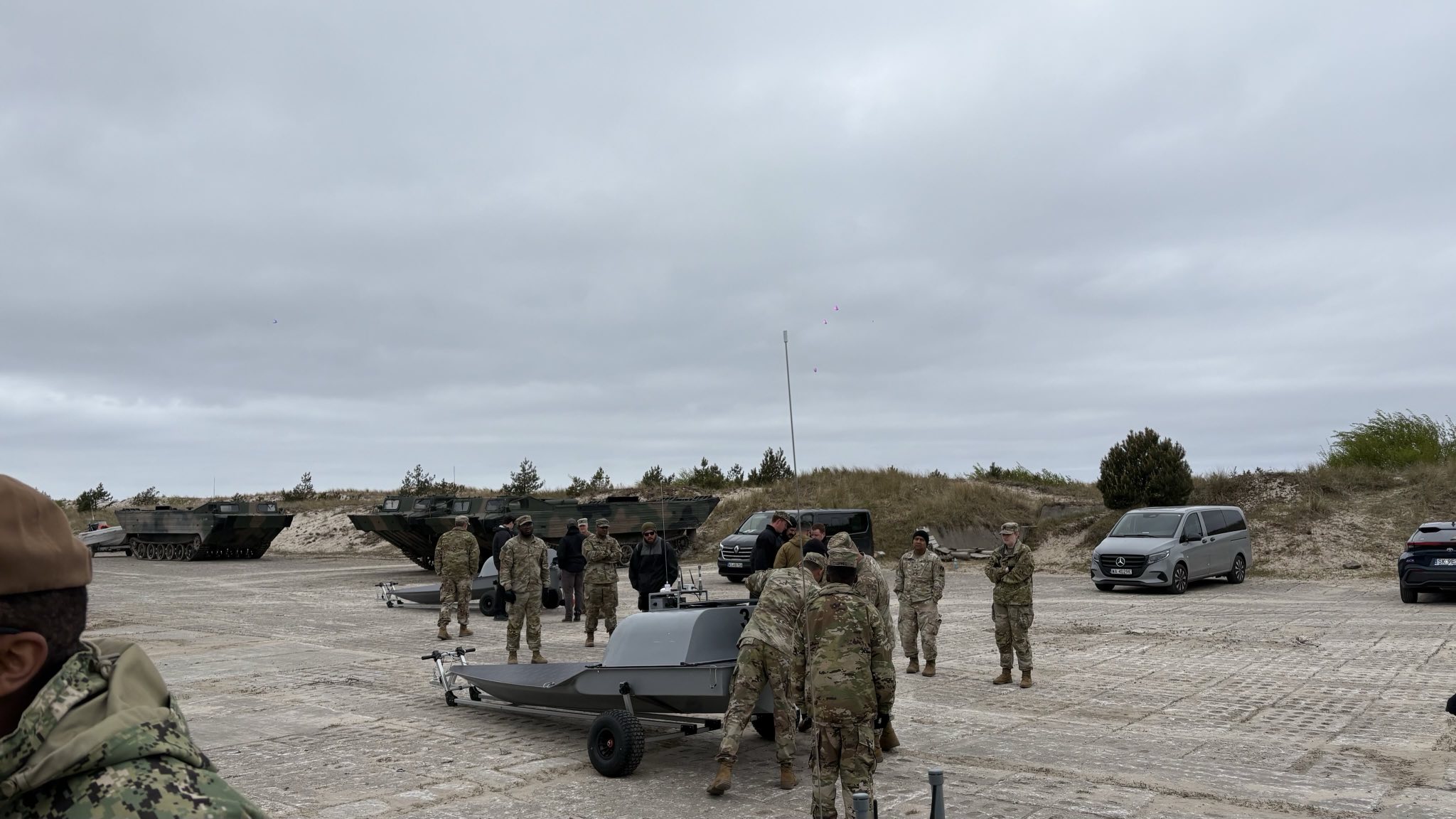 Several uniformed men and women from the US Army and Polish Armed Forces examining an Unmanned Naval Tech on a trailer outside.  Parked cars and army carrier vehicles in the background.