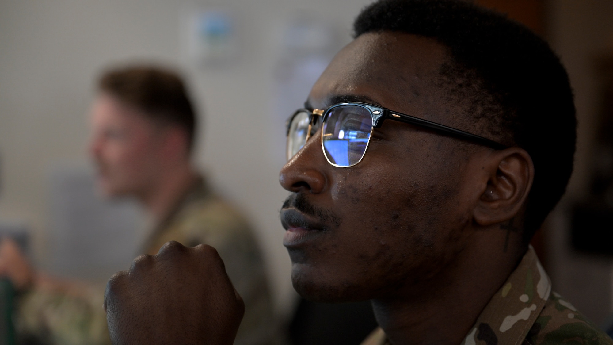 A U.S. Air Force service member in uniform and glasses sits at a workstation, looking intently at a monitor off-frame. Another uniformed individual is blurred in the background.