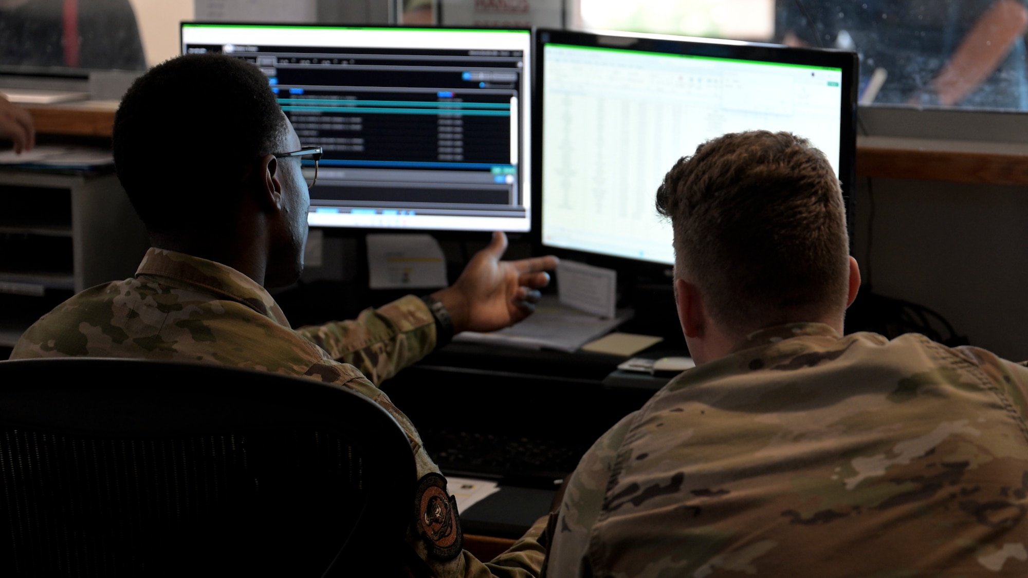 Two U.S. Air Force personnel in uniform sit side by side at a desk, viewing multiple computer monitors. One airman points at a screen displaying digital records, while the other observes.