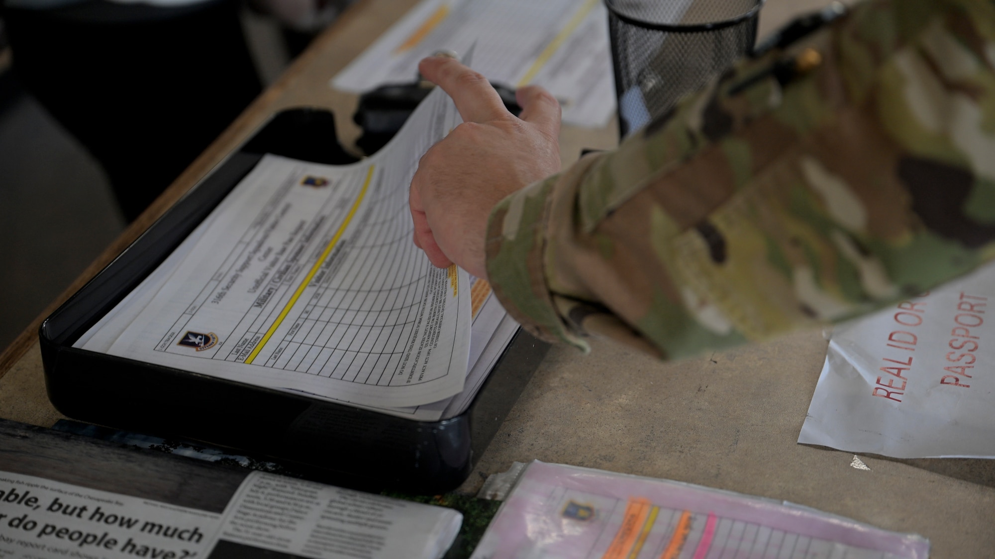 A person in a U.S. Air Force uniform fills out a visitor request form on a clipboard at a counter. Several forms, pens, and informational flyers are spread across the surface.