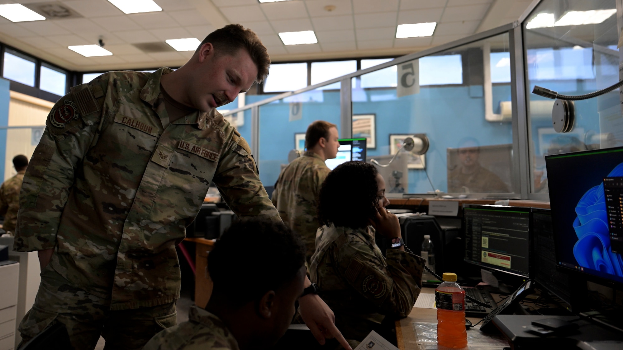 Four uniformed U.S. Air Force personnel are inside a visitor control center. One airman stands and points at a piece of paper held by a seated airman, while others sit at computer monitors. A wall of windows and security booths is visible in the background.
