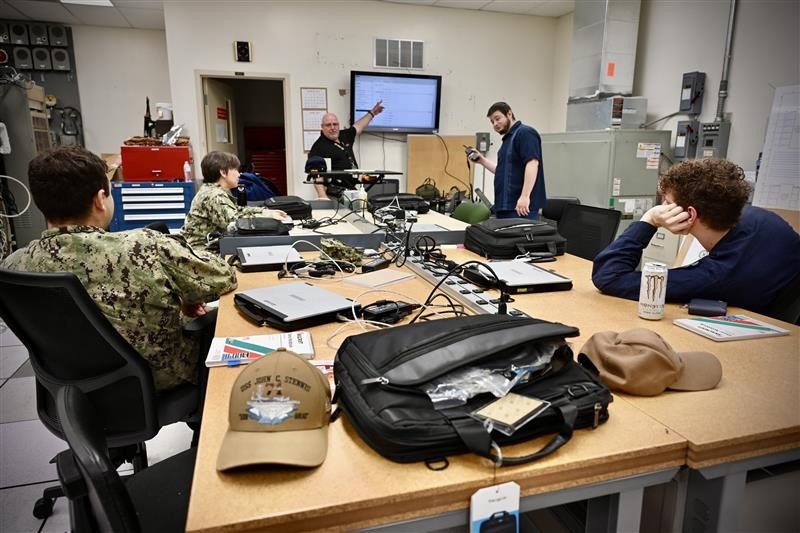A male and female Navy sailors with several NIWC Employees sitting at a table watching a man in a black polo shirt pointing at a display mounted to a wall.