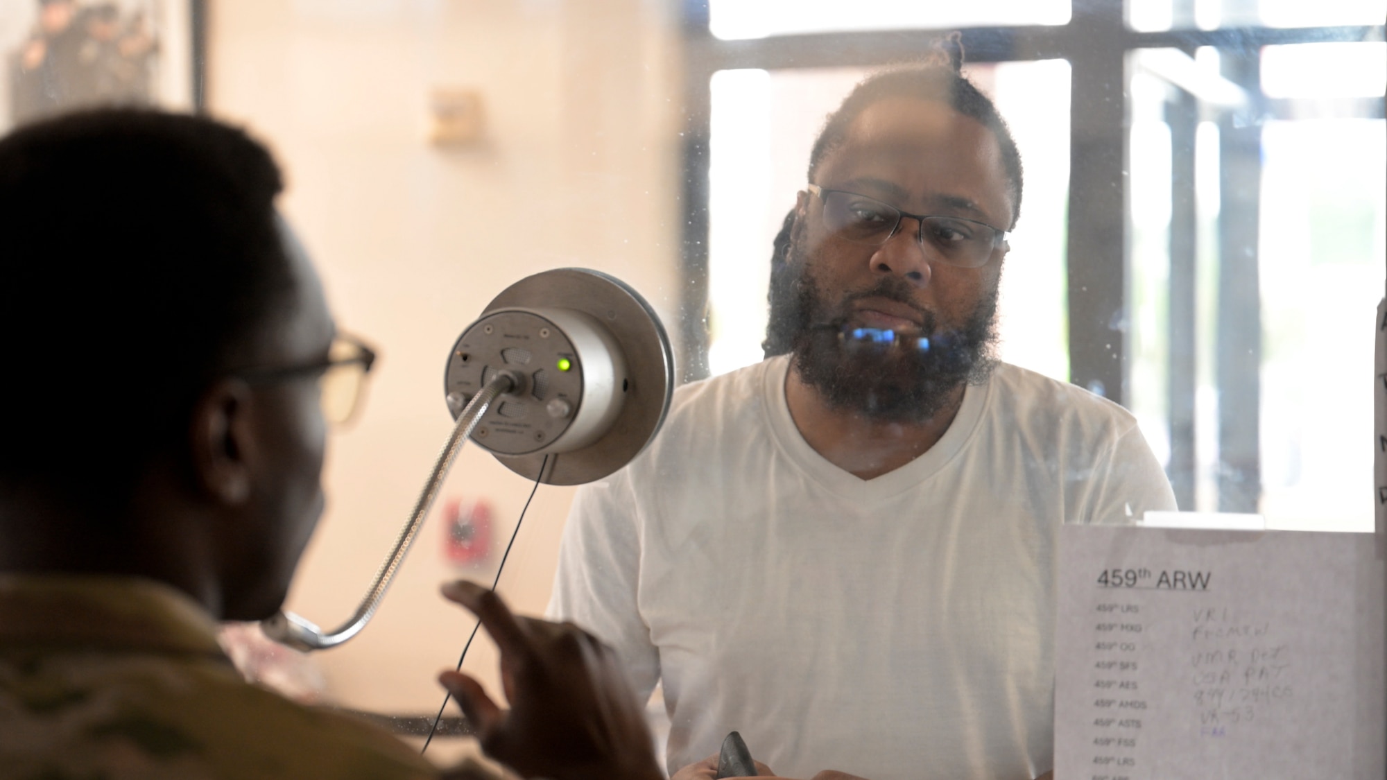 Senior Airman Marcellis Gillis of the U.S. Air Force, serving as a visitor control center clerk with the 316th Security Support Squadron, assists a male customer through a service window at the Visitor Control Center at Joint Base Andrews, Maryland, on July 21, 2025. A microphone and glass partition separate them. The center enforces security policies and verifies credentials. (U.S. Air Force photo by Staff Sgt. Isaac March)