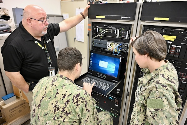 A male and female Navy sailors looking at a computer terminal with a NIWC Atlantic employee standing behind in a black polo shirt .