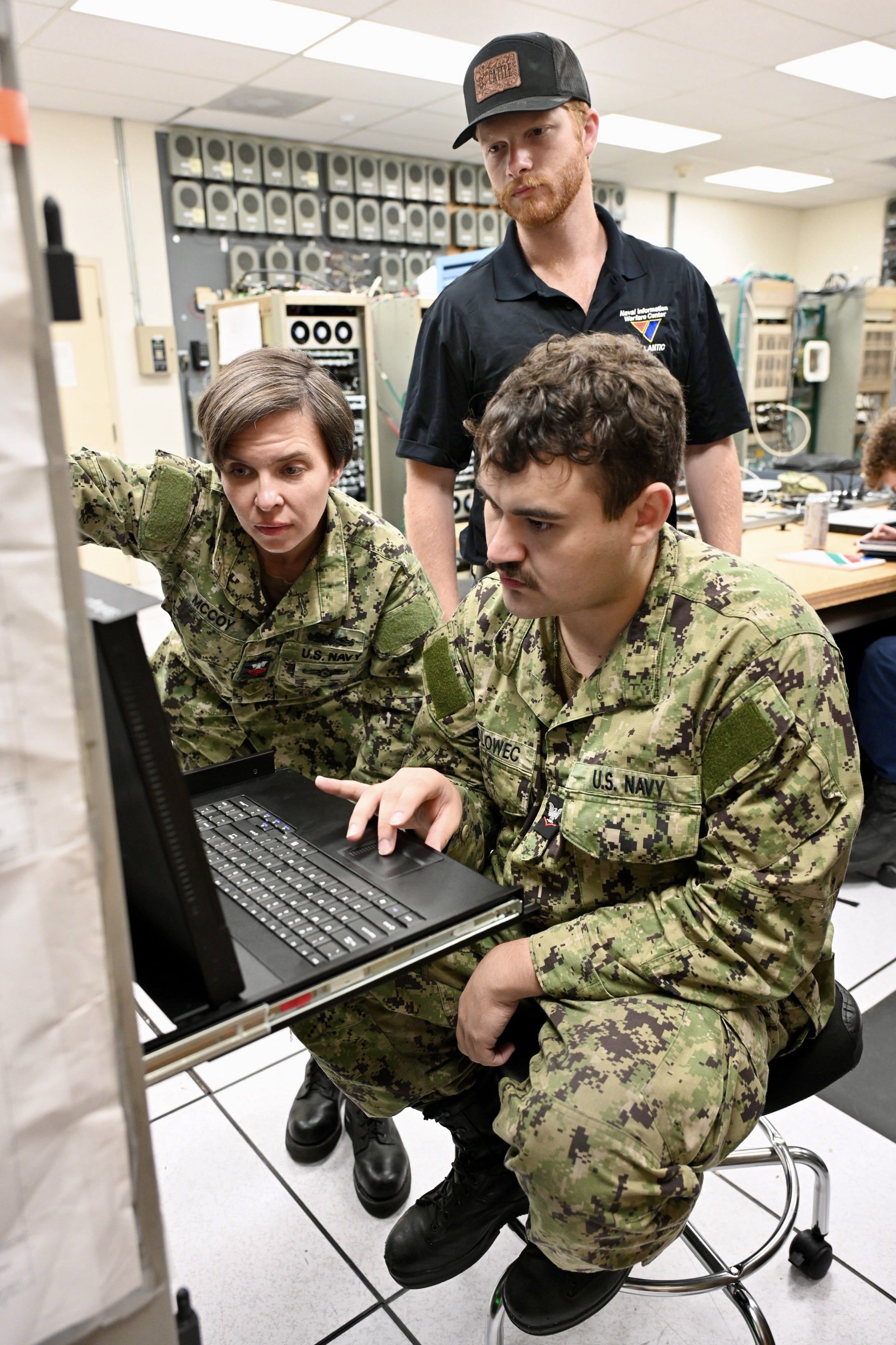 A male and female Navy sailors looking at a computer terminal with a NIWC Atlantic employee standing behind in a black polo shirt and black trucker cap.