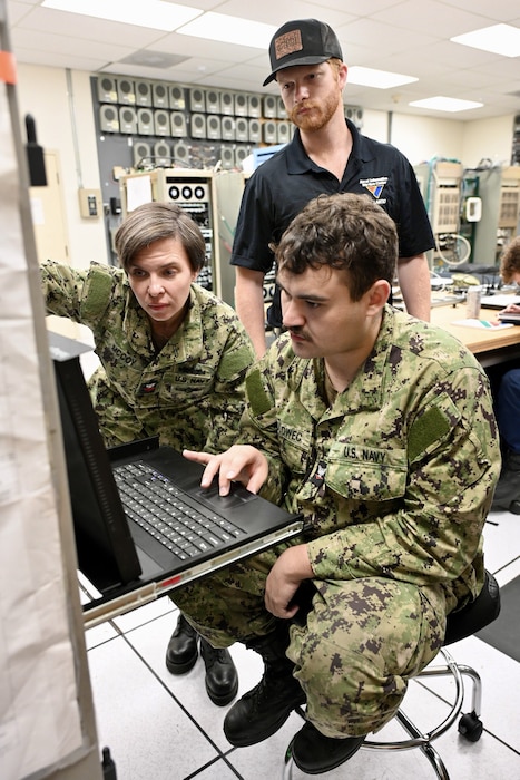 A male and female Navy sailors looking at a computer terminal with a NIWC Atlantic employee standing behind in a black polo shirt and black trucker cap.