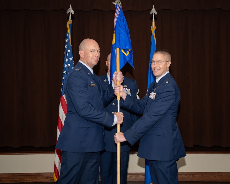 Two military members in blues service dress uniforms pose for a photo while holding the blue unit guidon