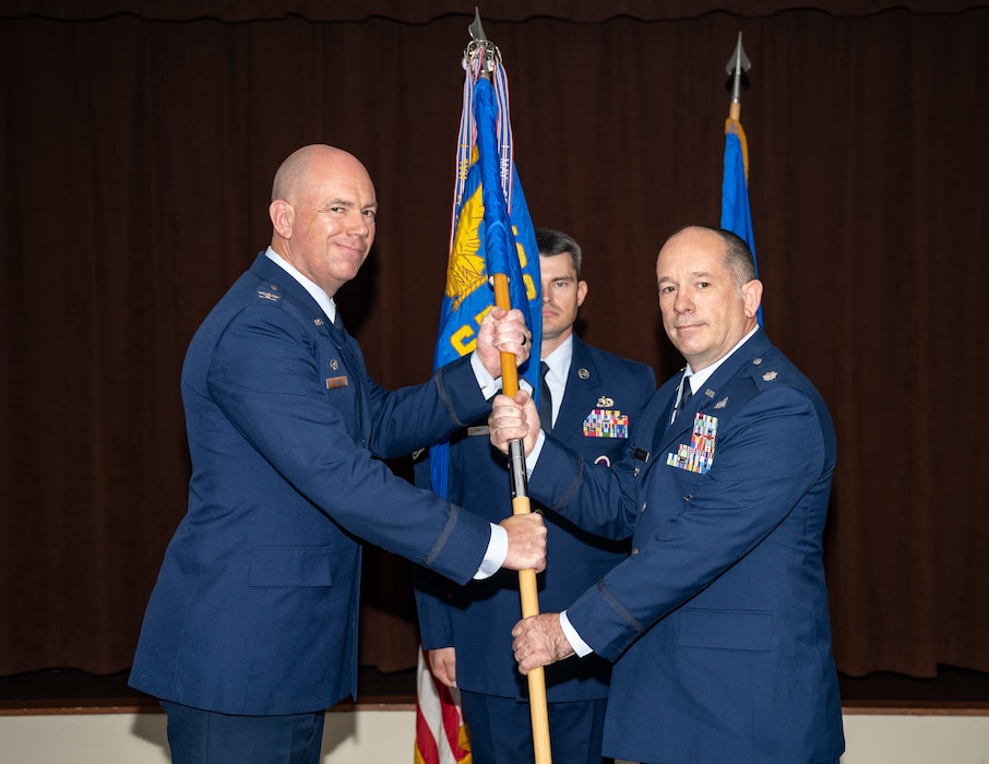 Two military members in blues service dress uniforms pose for a photo while holding the blue unit guidon