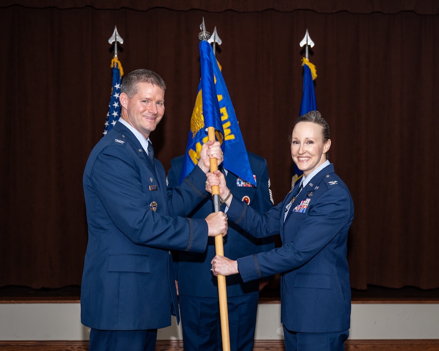 Two military members in blues service dress uniforms pose for a photo while holding the blue unit guidon