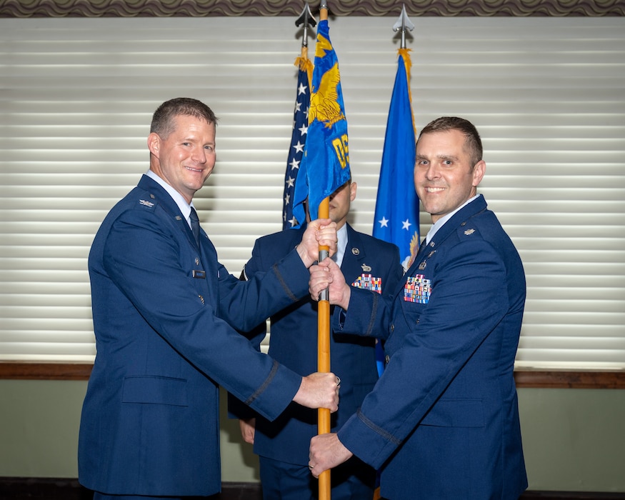 Two military members in blues service dress uniforms pose for a photo while holding the blue unit guidon