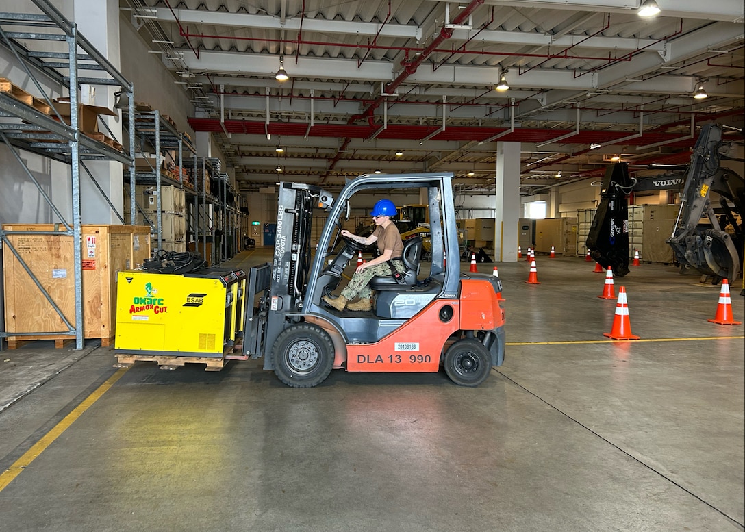A person sits in the drivers seat of an orange forklift inside a large warehouse.