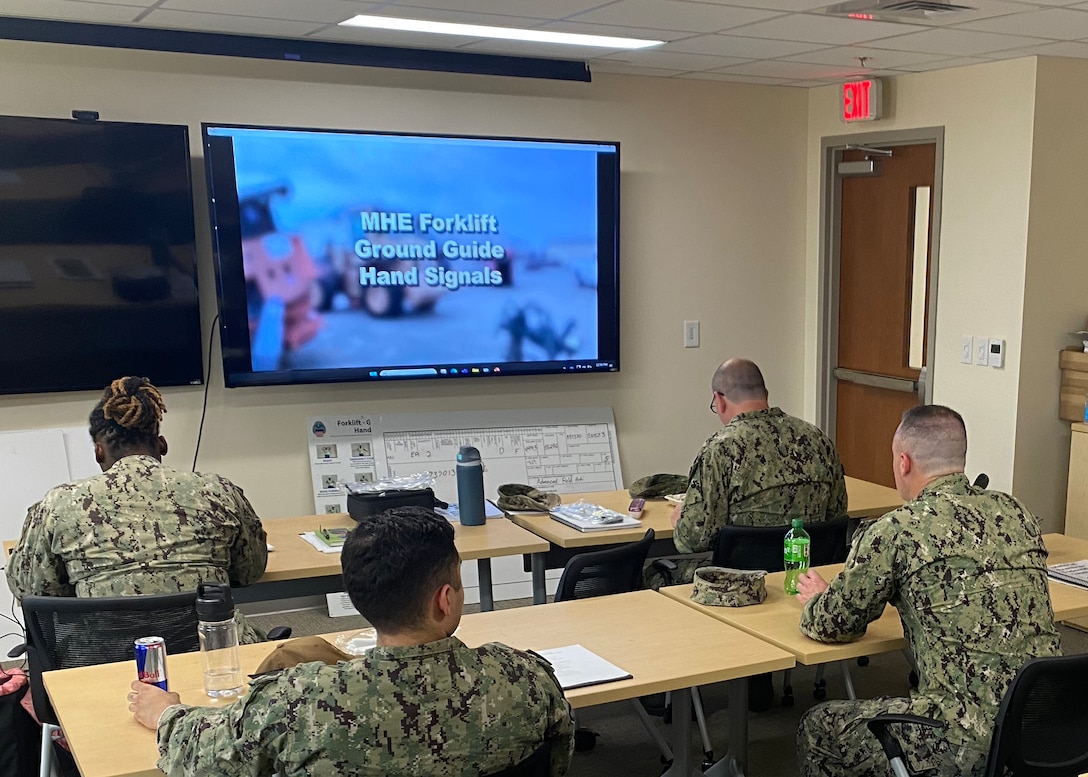 Four men dressed in military working uniforms sit in a classroom looking towards a screen. The screen has a training lesson playing.