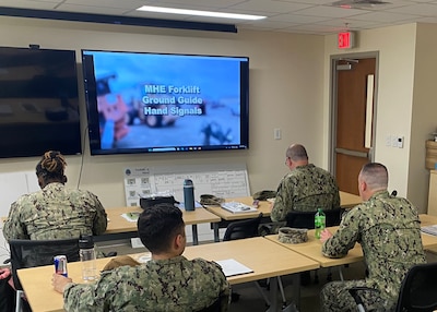 Four men dressed in military working uniforms sit in a classroom looking towards a screen. The screen has a training lesson playing.