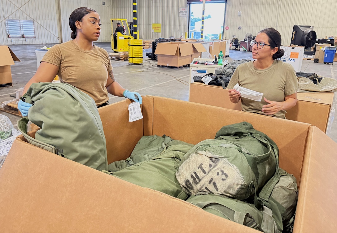 Two women dressed in military uniforms stand in a warehouse next to a large box with military duffel bags inside it.