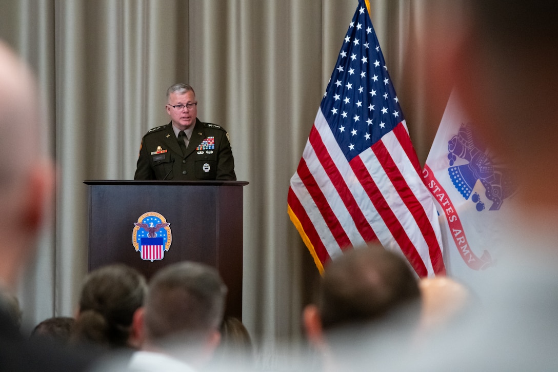 A man wearing a military uniform stands on stage behind a lectern speaking to a crowd.