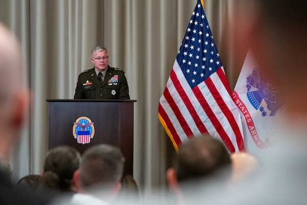 A man wearing a military uniform stands on stage behind a lectern speaking to a crowd.